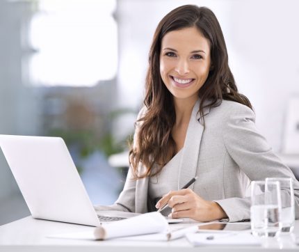 Eine Frau sitzt lächelnd an ihrem Arbeitsplatz hinter ihrem Laptop und ihren Notizen.